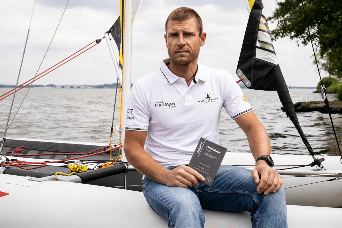 Natural Pharmaceuticals ProMan® Plus Żeglarz w białej koszulce polo Team ProMan® Plus siedzi na jachcie, trzymając opakowanie suplementu diety ProMan® Plus.