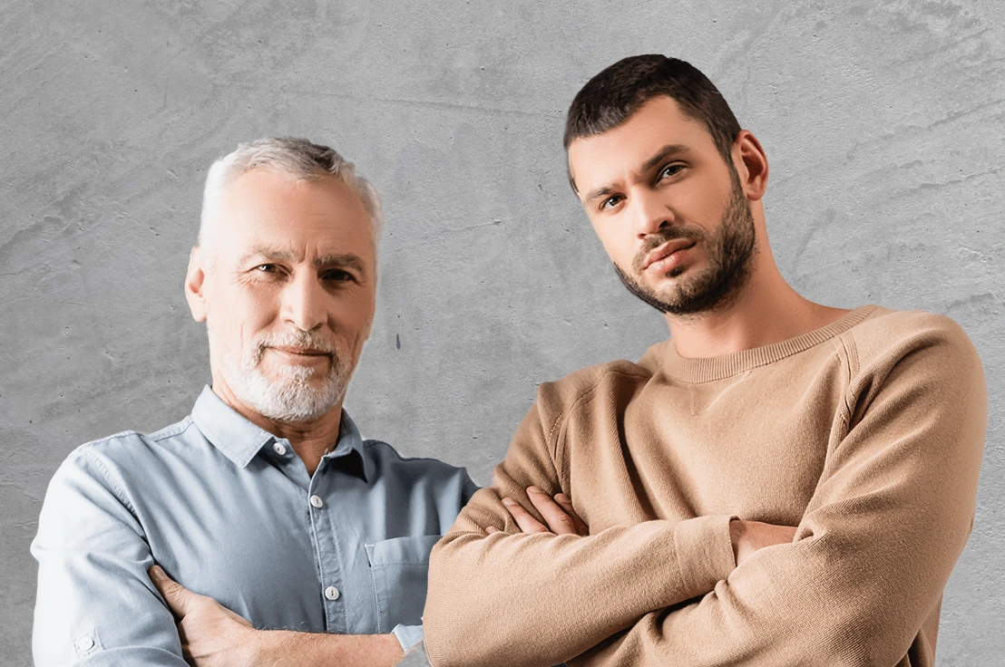 Natural Pharmaceuticals ProMan® Plus Dwóch mężczyzn - starszy z siwymi włosami i brodą oraz młodszy z ciemną brodą - stoją z założonymi rękami na szarym tle.