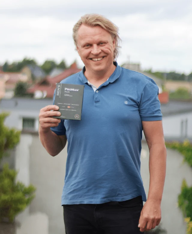 Natural Pharmaceuticals ProMan® Plus Uśmiechnięty mężczyzna z jasnymi włosami w niebieskiej koszulce polo, trzymający opakowanie ProMan® Plus na tle panoramy miasta.