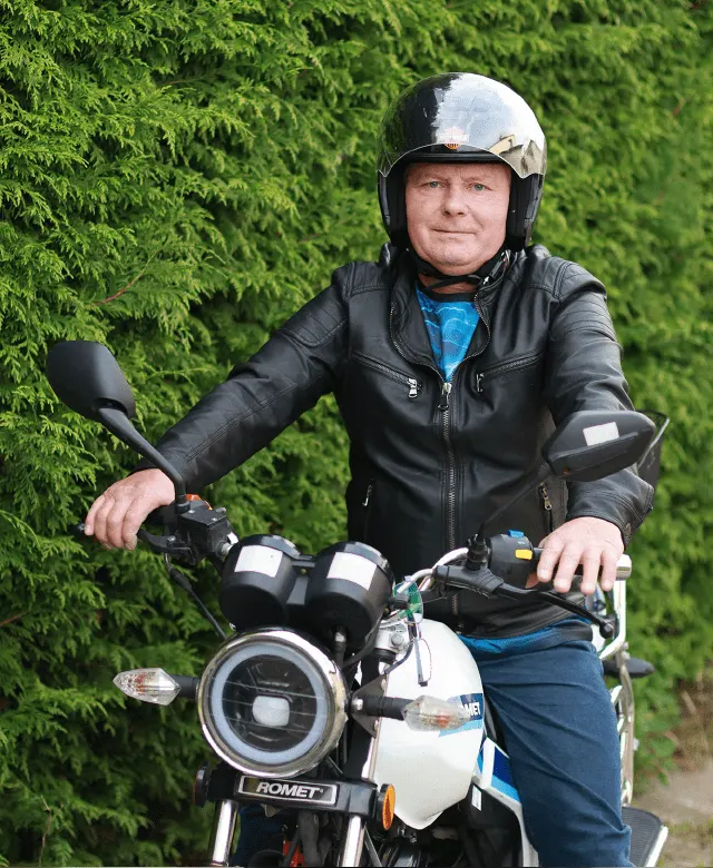 Natural Pharmaceuticals ProMan® Plus Mężczyzna w skórzanej kurtce i kasku motocyklowym, siedzący na motorze Romet na tle zielonych tui.